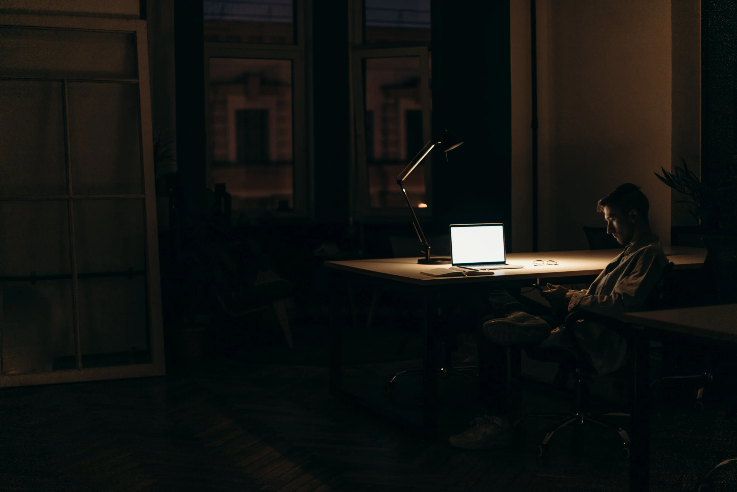 A person sits in a dimly lit room, working on a laptop at a desk.