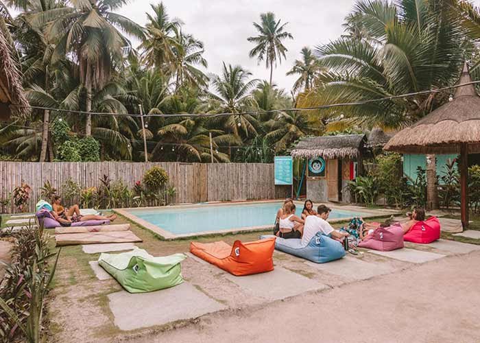 People relax on colorful bean bags around a small outdoor pool surrounded by palm trees and thatched-roof huts.