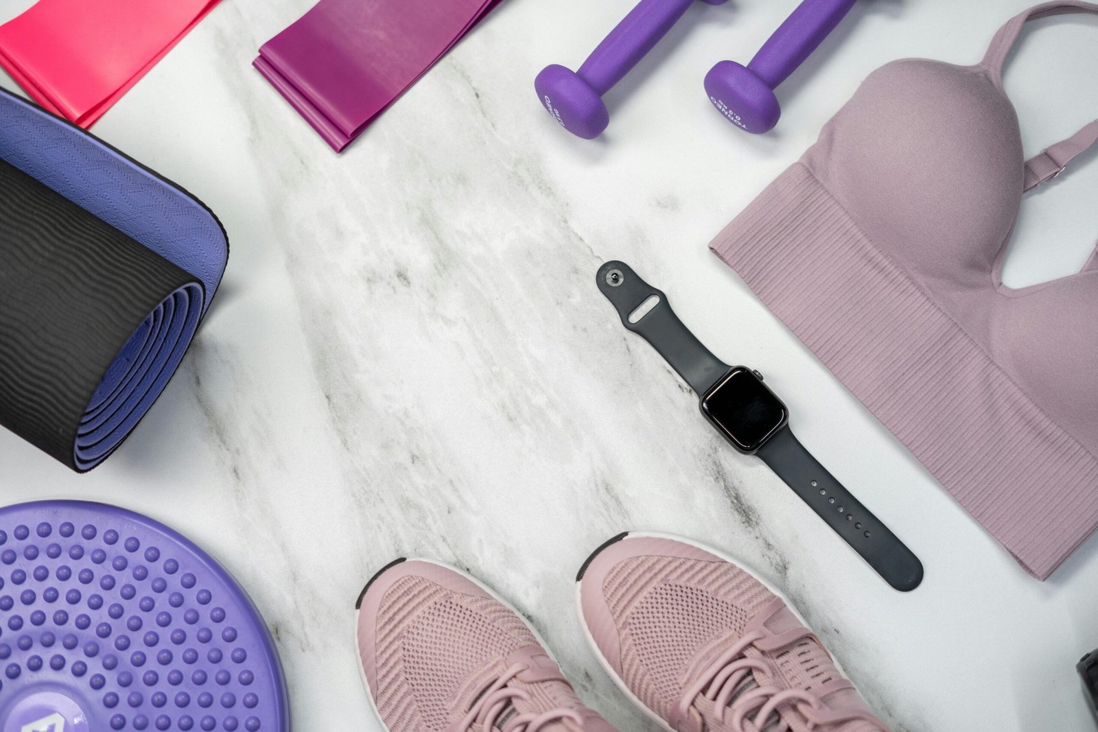 Flat lay of fitness equipment including a yoga mat, resistance bands, dumbbells, sports bra, smartwatch, balance disk, and athletic shoes on a marble surface.