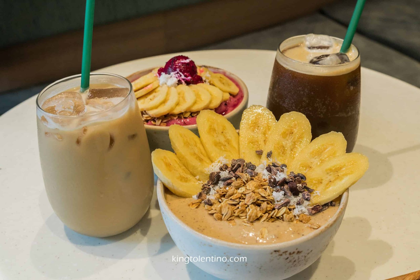 Two glasses of iced coffee and two smoothie bowls topped with banana slices and granola are arranged on a round table.
