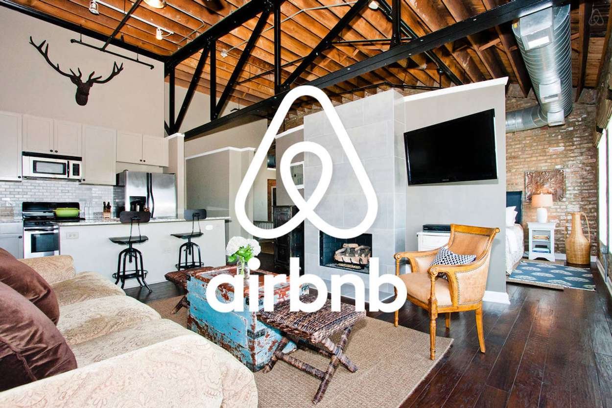 Loft-style living room with exposed beams, modern kitchen, mounted TV, and a bedroom in the background; Airbnb logo overlayed at center.