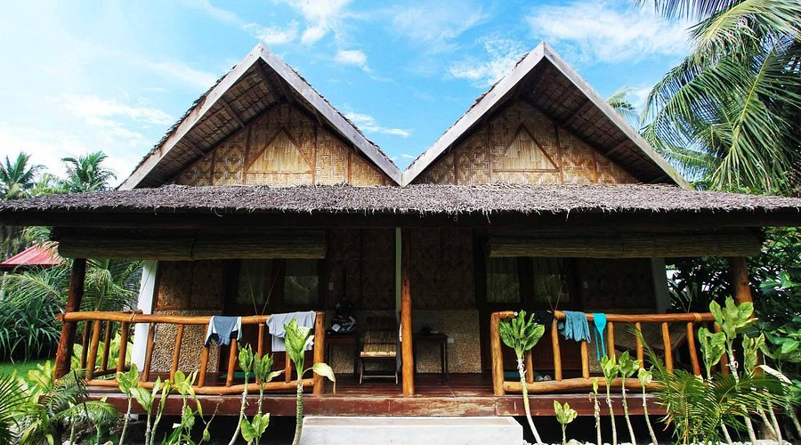 A traditional bamboo cottage with a thatched roof, two peaked sections, a wooden porch, and clothes hanging over the railing, surrounded by greenery and palm trees.