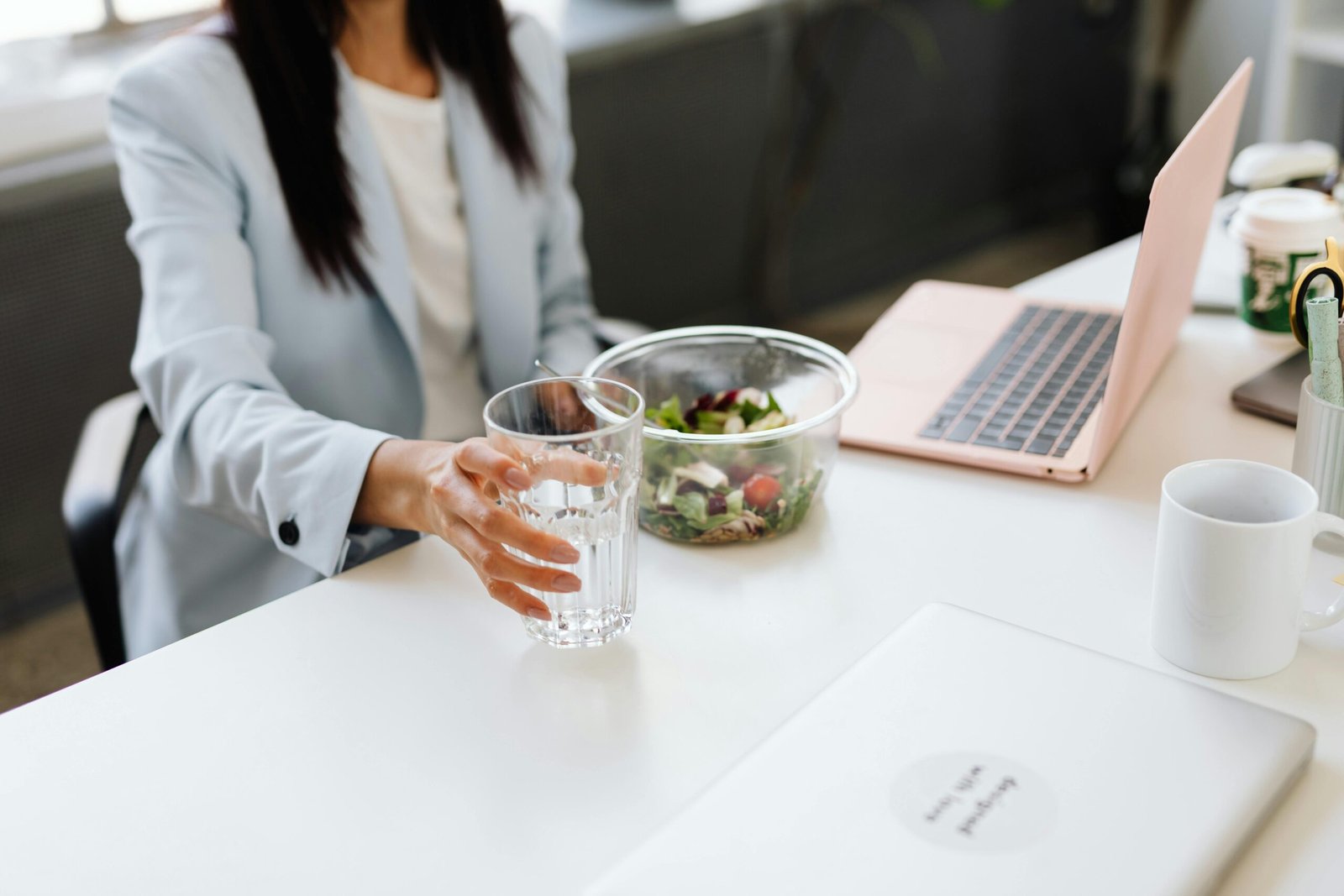 A person in business attire sits at a desk with a laptop, salad bowl, and white mug, reaching for a glass of water.