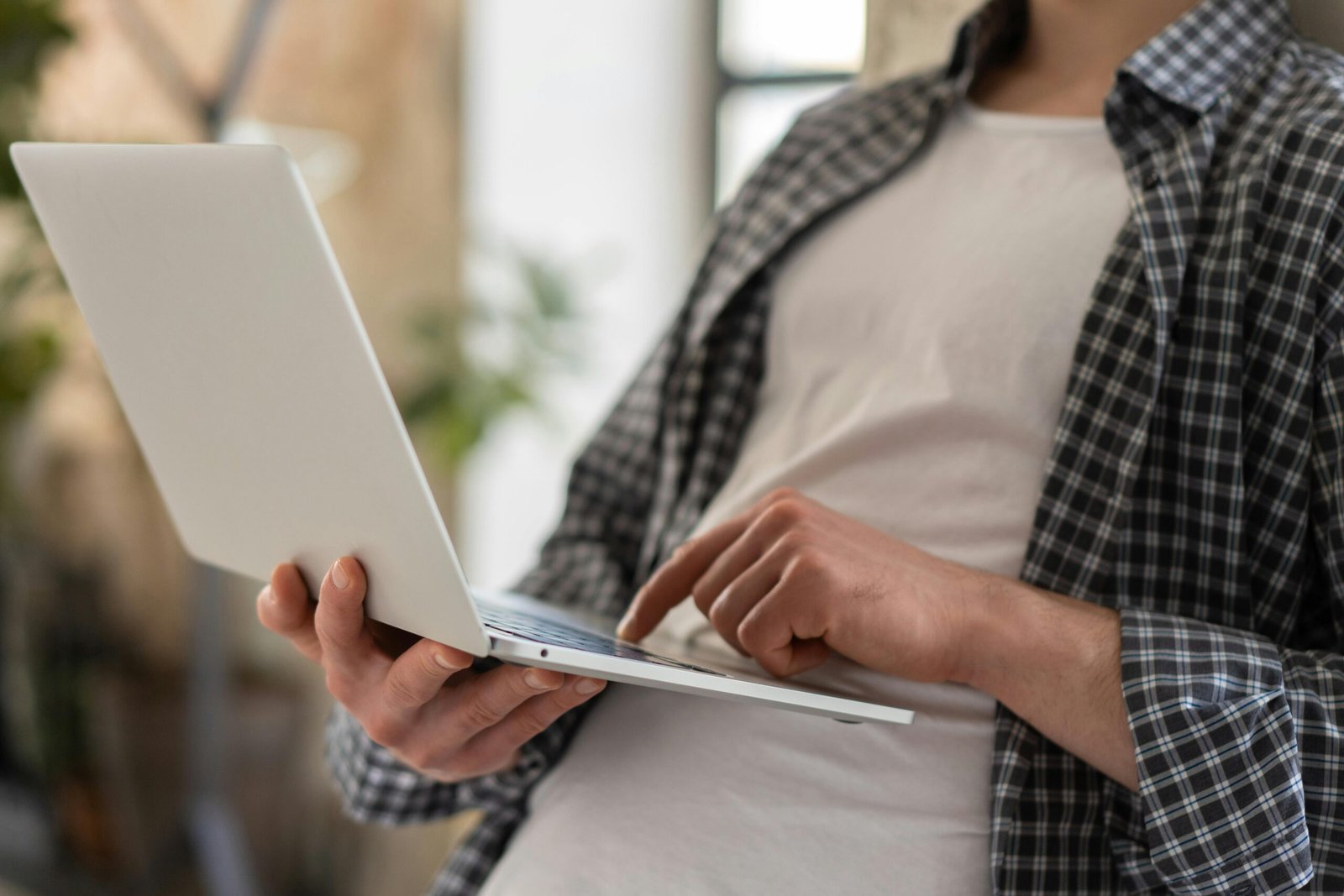 A person wearing a plaid shirt and white t-shirt is using a laptop, holding it with one hand and typing with the other.