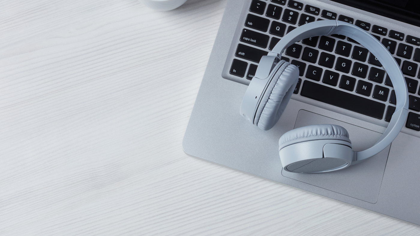 A pair of wireless gray headphones rests on the keyboard of an open silver laptop on a light wooden surface.
