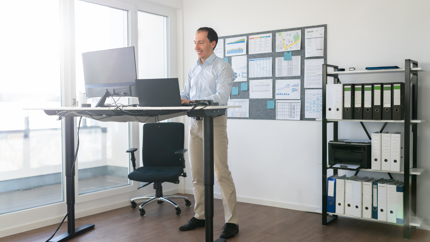 A man stands at a height-adjustable desk working on a laptop in a bright office with charts on the wall and shelves of binders. A man stands at a height-adjustable desk working on a laptop in a bright office with charts on the wall and shelves of binders.