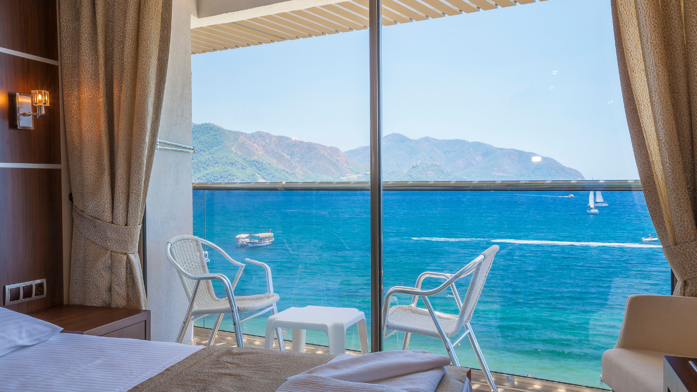 Hotel room with a bed and chairs on a balcony overlooking the sea, with mountains and boats visible in the distance through large glass doors. Hotel room with a bed and chairs on a balcony overlooking the sea, with mountains and boats visible in the distance through large glass doors.