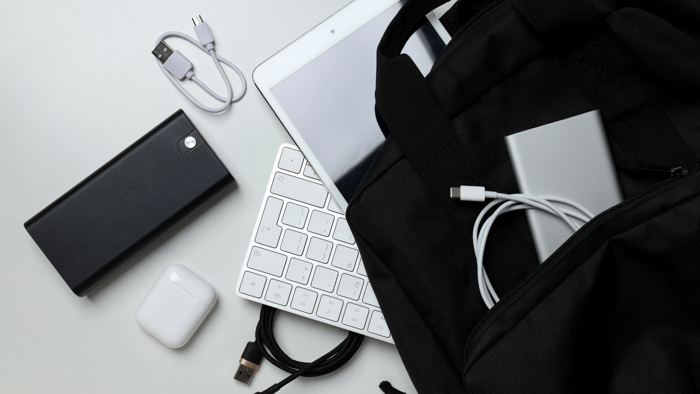 A black backpack, white tablet, wireless keyboard, headphones case, power bank, portable drive, and charging cables arranged on a white surface. A black backpack, white tablet, wireless keyboard, headphones case, power bank, portable drive, and charging cables arranged on a white surface.