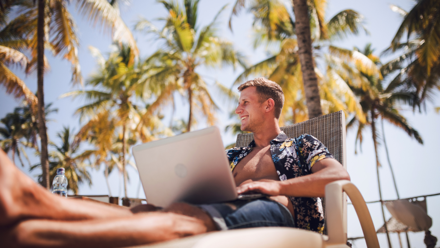 A man sits in a lounge chair outdoors with a laptop on his lap, surrounded by palm trees on a sunny day.
